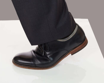 Black dress shoe with brown sole worn with black pants on a white platform.